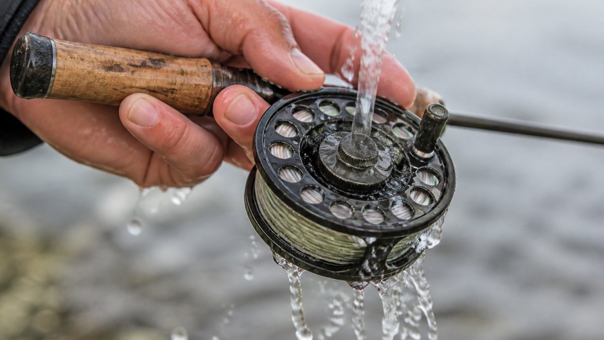 Close-up of a fly reel being rinsed with fresh water after saltwater fishing, showing water droplets cascading over the sealed drag mechanism and spool