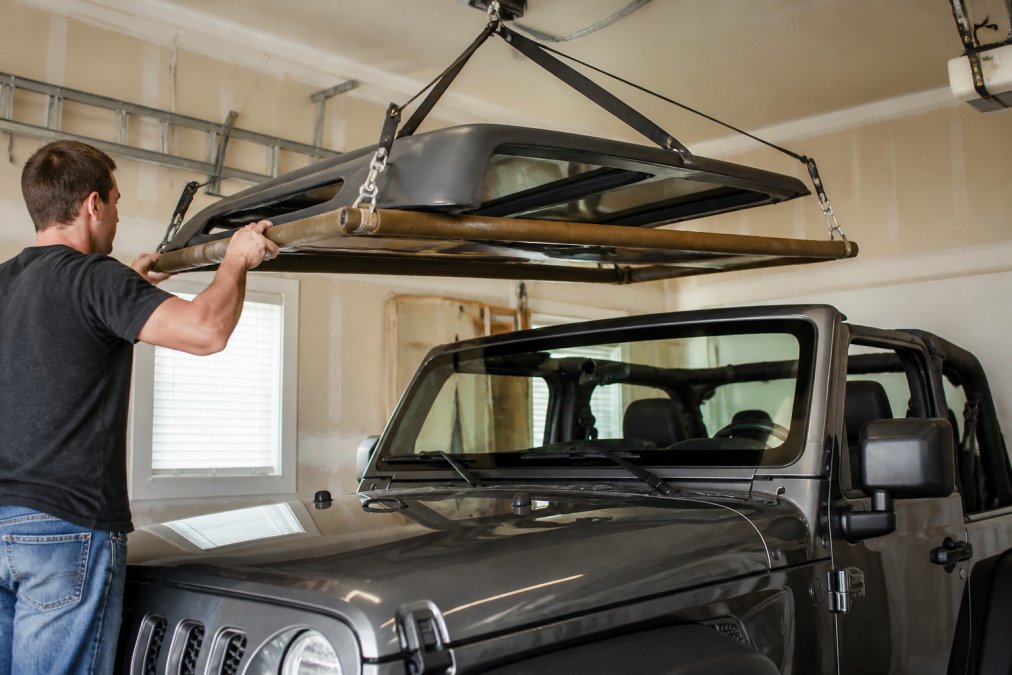 A Jeep Wrangler owner in a residential garage carefully guiding a hardtop onto a padded hoist platform, with the hardtop suspended above the vehicle hood and safety straps visible