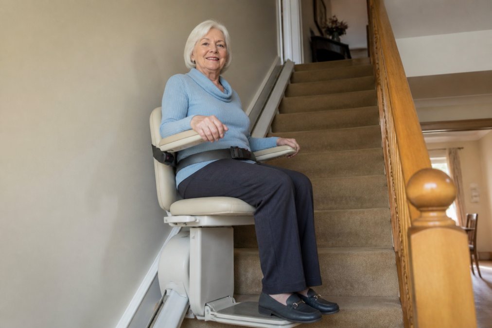 Older adult safely seated in a stairlift with visible safety features including padded armrests, secure seat belt, and footrest support, positioned mid-staircase in a residential home with warm natural lighting