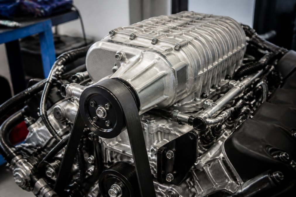 Close-up of a twin-screw supercharger assembly mounted directly on an engine block, showing precision aluminum components, belt drive system, and bolt-on fitment points under bright workshop lighting