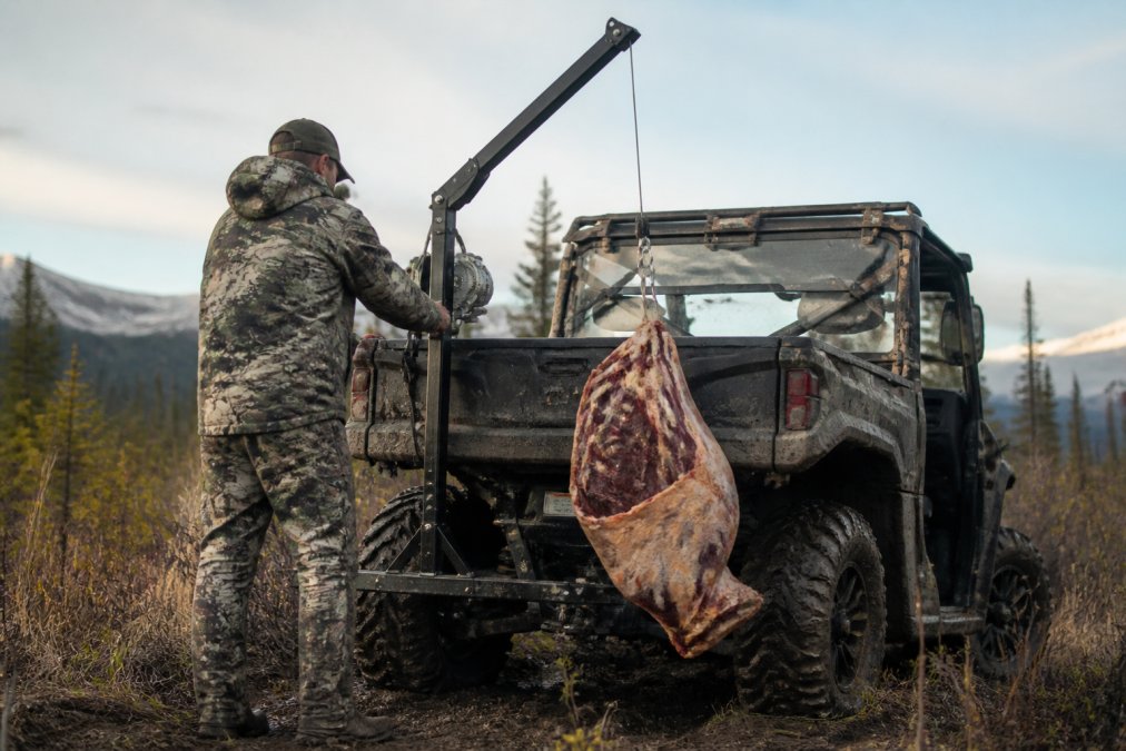 Hunter using a hitch-mounted UTV game hoist to lift a moose quarter in rugged, muddy bush terrain.