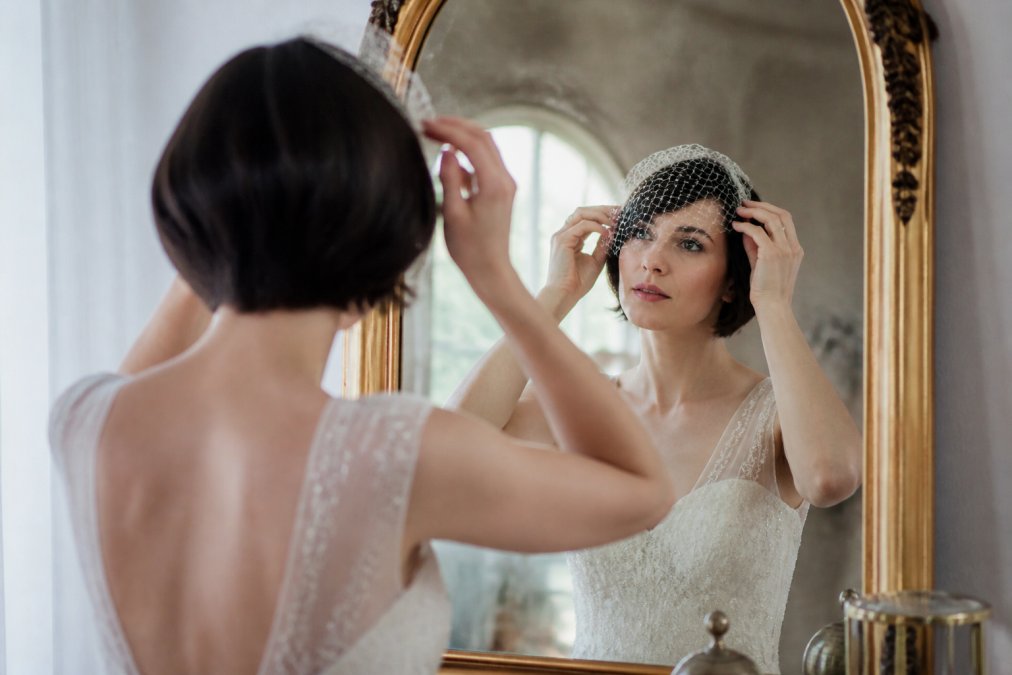 A bride with a short bob hairstyle adjusting a vintage birdcage veil while looking into a sunlit mirror.