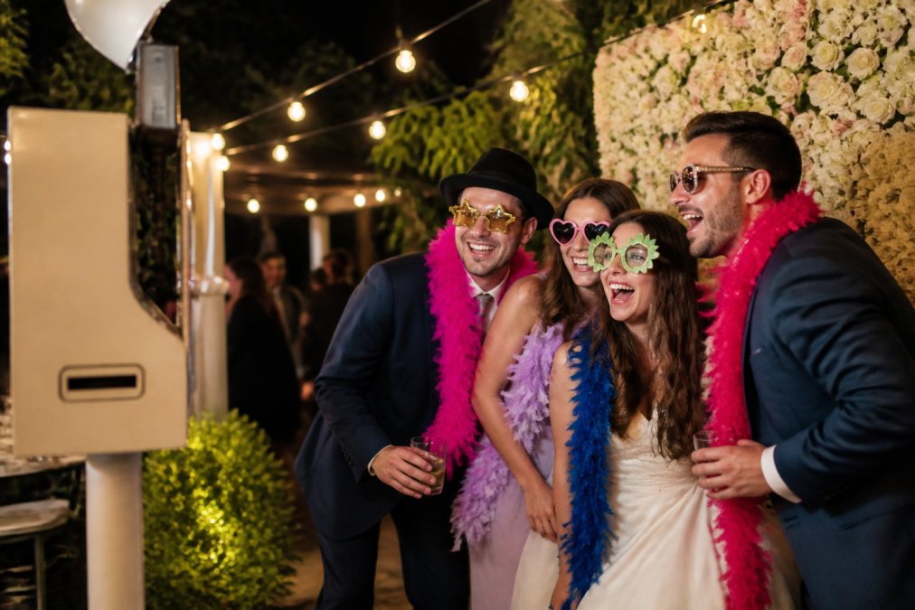 Guests laughing and posing with feather boas and novelty glasses inside an open-air photo booth at a wedding reception, string lights and a floral backdrop glowing in the background