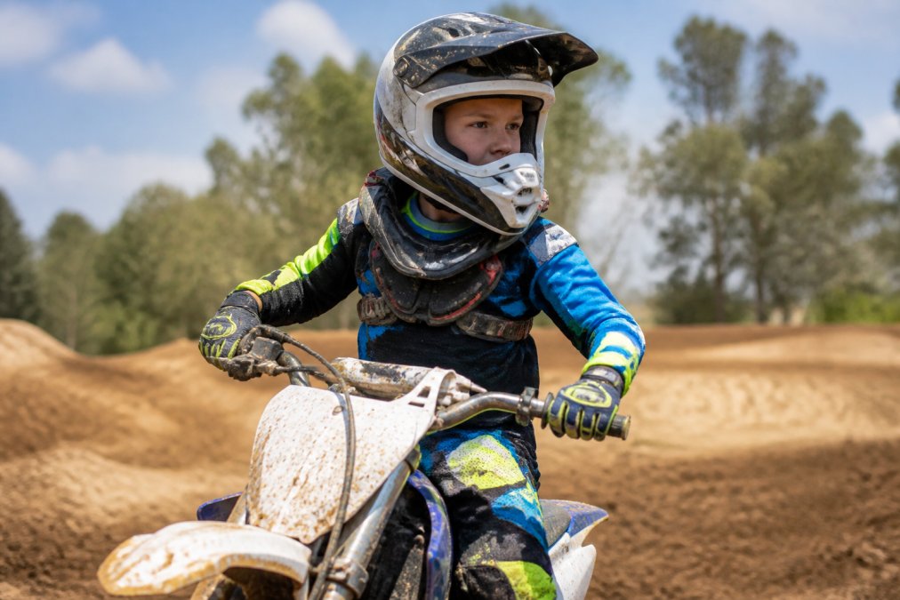 Young motocross rider in full protective gear on dirt bike at outdoor training facility during daytime, demonstrating proper safety equipment positioning during active riding