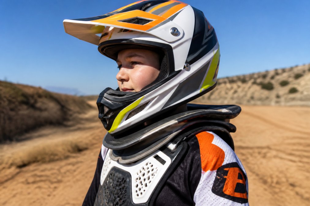 Child motocross rider wearing properly fitted neck brace with visible collarbone clearance, helmet positioned correctly, and chest protector aligned, shown from side angle in bright daylight