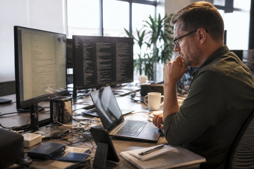 Business professional troubleshooting system issues at desk with multiple monitors displaying automation dashboards and technical documentation in modern office setting