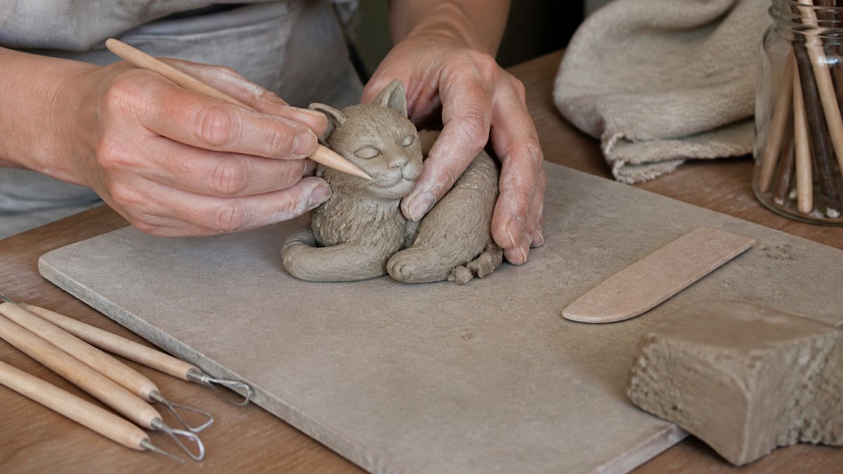 Hands shaping a small air-dry clay sculpture on a ceramic tile work surface, showing malleable texture and detail work with sculpting tools nearby