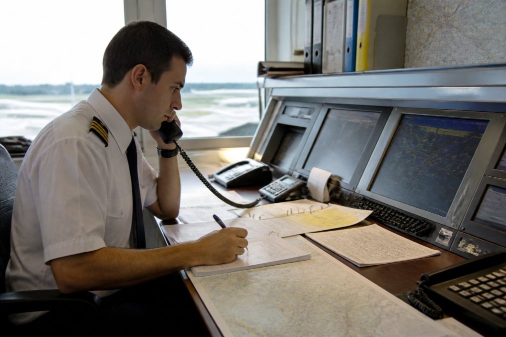 Aircraft dispatcher at a dispatch desk monitoring multiple radar screens and communication equipment, wearing professional aviation attire, with flight data displays and weather maps visible in background lighting