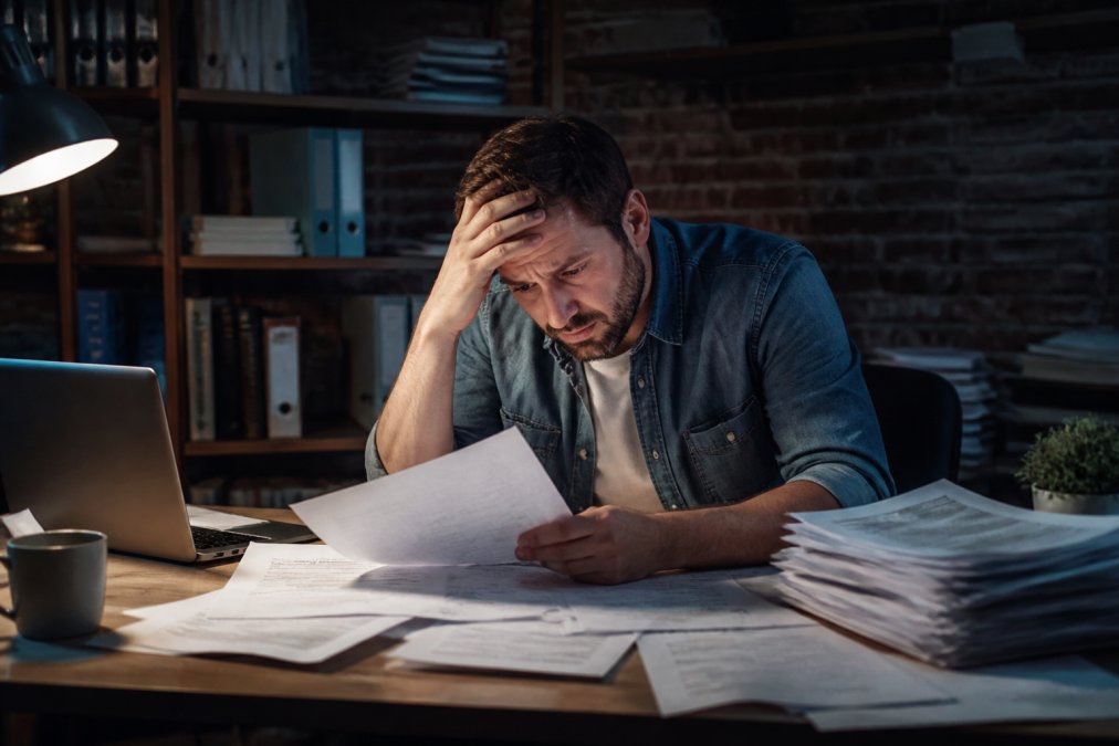 Is All in One Software Worth It? A 2026 Cost Analysis A small business owner at a cluttered desk with multiple browser tabs open showing different software dashboards, looking overwhelmed while reviewing subscription invoices on paper