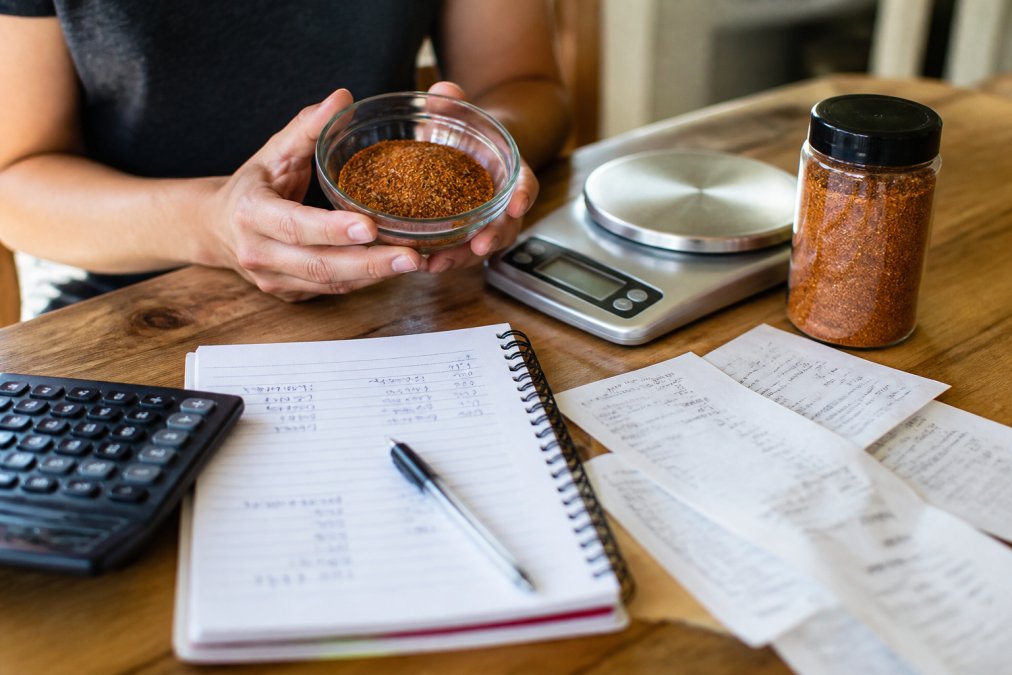 Close-up of hands holding a small bowl of dry rub next to a kitchen scale and a jar of store-bought rub on a wooden countertop