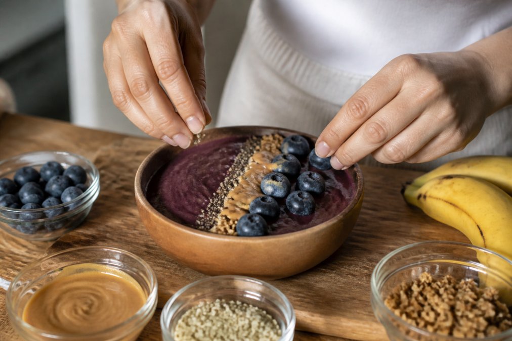 Close-up of a person's hands carefully arranging hemp seeds, a thin almond butter drizzle, and fresh blueberries onto a thick, dark purple acai bowl base in a clean, well-lit kitchen, wooden countertop in the background