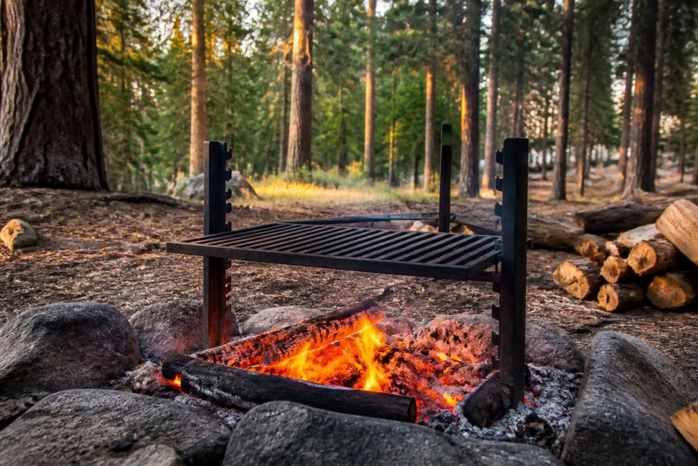 A heavy-duty carbon steel campfire grill positioned over a glowing open wood fire at a forest campsite, with the grate raised to mid-height above orange embers, surrounded by tall pines in late afternoon light