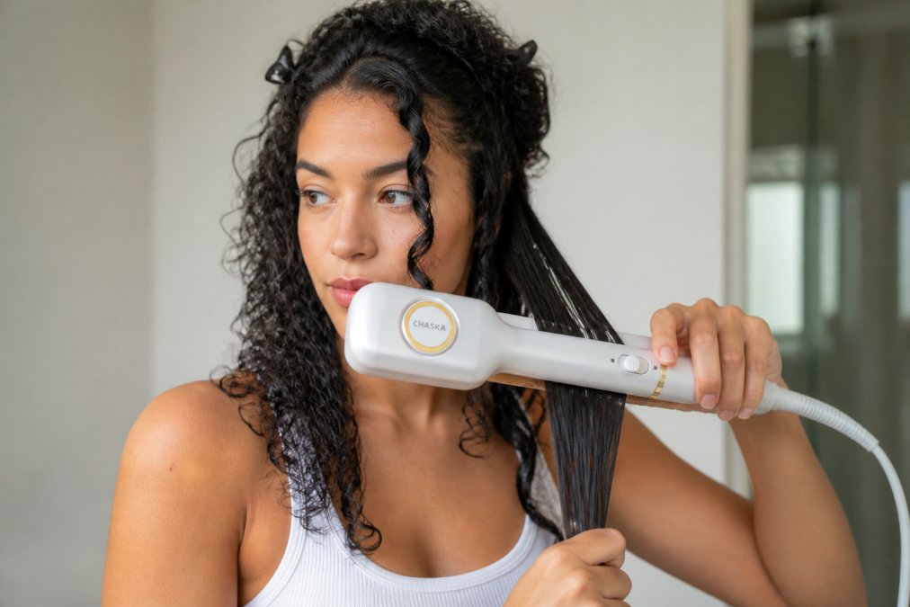 Woman with thick, curly hair using an all-in-one styler to straighten sections, showing the styling motion and technique on damp hair with natural lighting