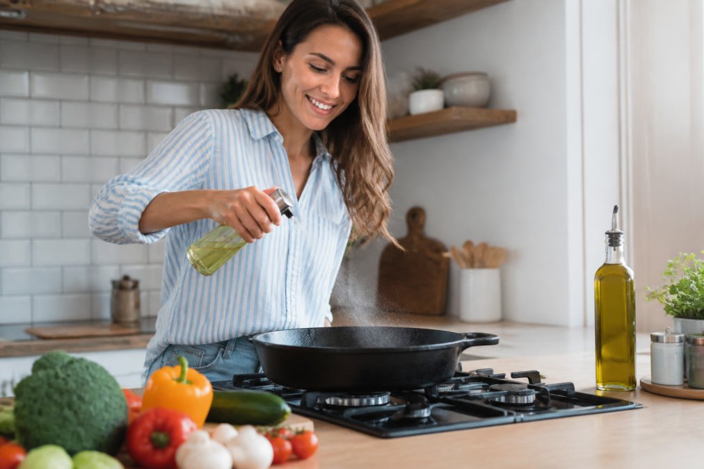 A home cook in a bright kitchen using a glass oil sprayer to mist a cast iron skillet, with fresh vegetables and a bottle of olive oil on the counter nearby