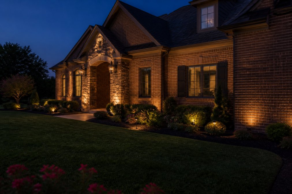 A brick home exterior at dusk with subtle warm uplighting along the foundation illuminating the texture of the masonry, accent lighting on architectural details like a stone entryway, and strategic landscape lighting creating depth without harsh shadows or artificial brightness