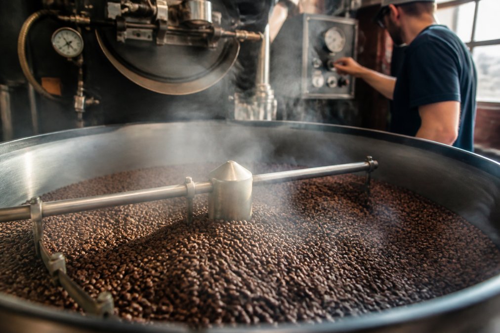 Close-up of freshly roasted coffee beans cooling on a metal roasting table, showing rich brown and dark chocolate colors with visible steam rising, a professional roaster in background monitoring temperature gauges and adjusting controls