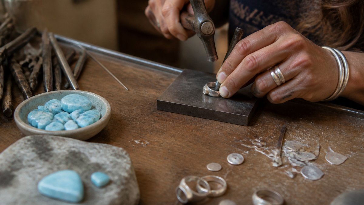 Close-up of a silversmith's hands working on a handmade sterling silver ring at a workbench, with hammering tools, metal scraps, and genuine Larimar stones visible in natural workshop lighting
