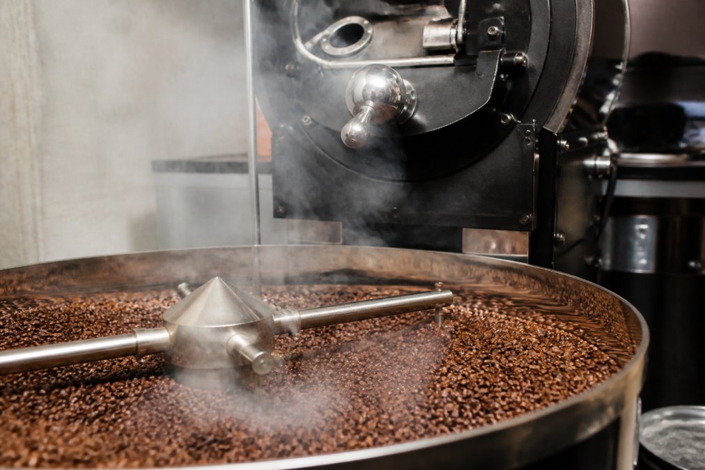 Close-up of freshly roasted coffee beans cooling in a drum roaster, showing rich brown color and steam rising, with roaster visible in background