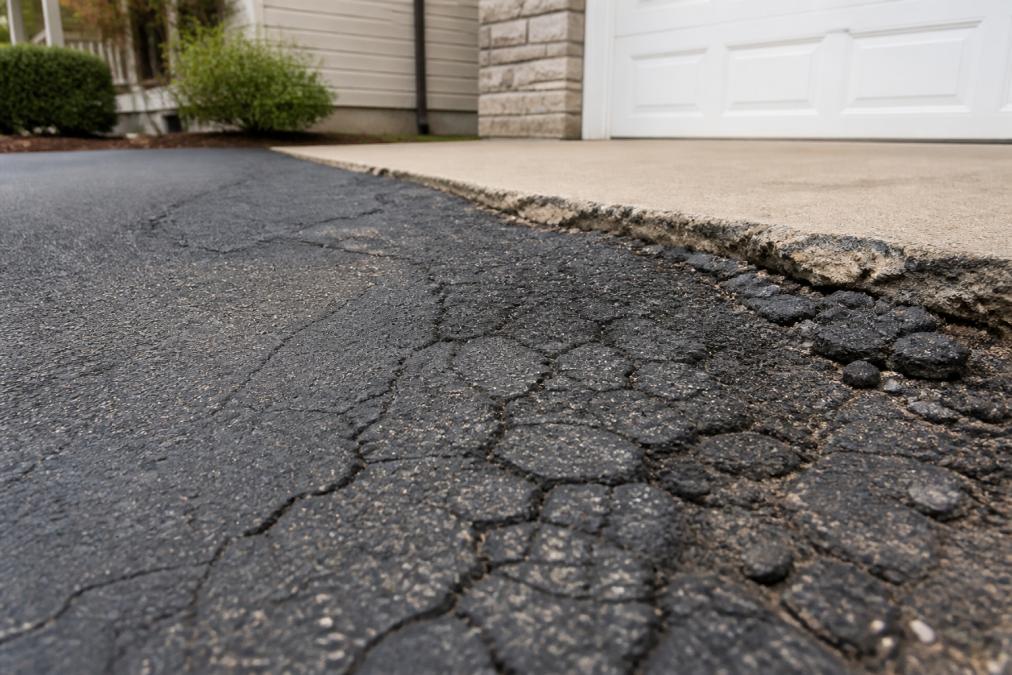 Close-up photograph of a residential asphalt driveway showing visible alligator cracking and crumbling edges near the garage apron, with a home exterior softly blurred in the background