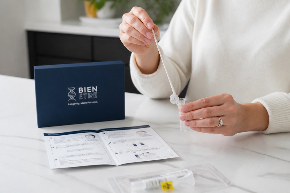 Person's hands performing a saliva swab collection at home, carefully placing the swab in a stabilization tube on a clean white countertop, with a collection kit and instructions visible
