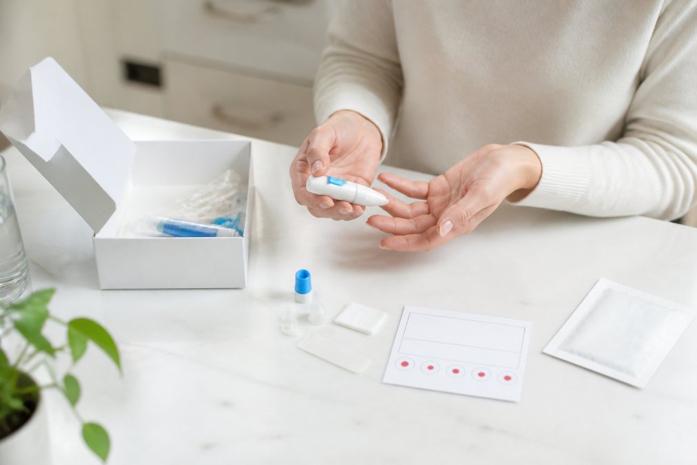 A person's hands preparing an at-home finger-prick blood sample kit on a clean kitchen table, with the test kit box, lancet, and collection card visible in soft daylight