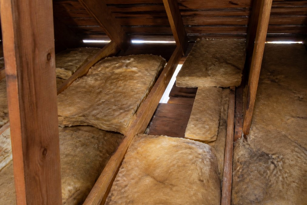 Close-up view of sparse, thin attic insulation showing exposed wooden rafters and visible gaps between fiberglass batts, demonstrating inadequate coverage with daylight visible through gaps