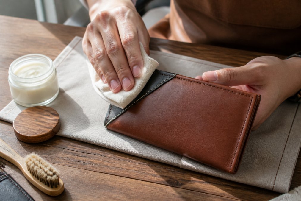Close-up of hands carefully applying leather conditioner to a genuine leather handbag using a soft cloth in gentle circular motions, with natural daylight illuminating the rich brown leather