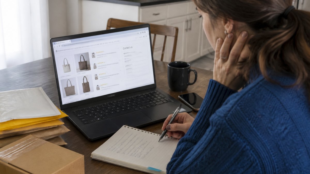A person at a kitchen table with a laptop, scanning an online store's reviews and contact page while taking notes on a notepad