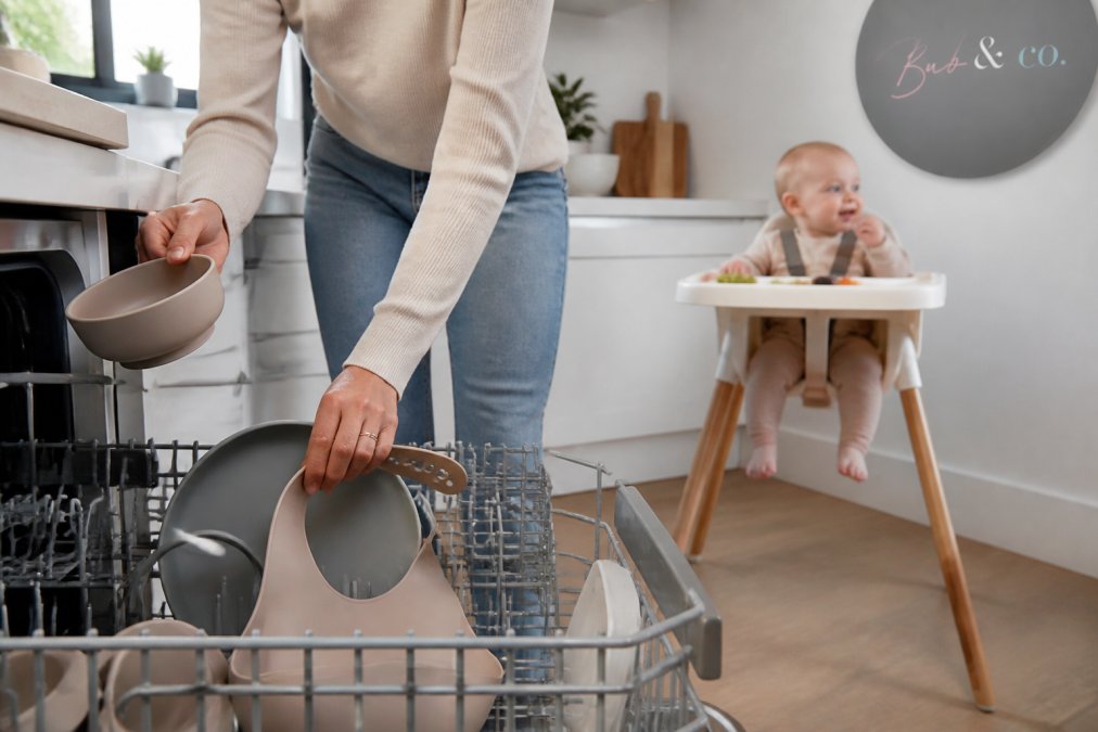 Parent loading silicone baby-led weaning bowls and bibs into a dishwasher while a baby eats in a highchair