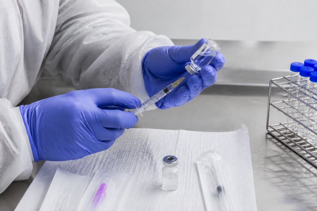 Hands performing aseptic technique during peptide reconstitution with sterile syringe, gauge needle, and bacteriostatic water vial on a clean laboratory surface