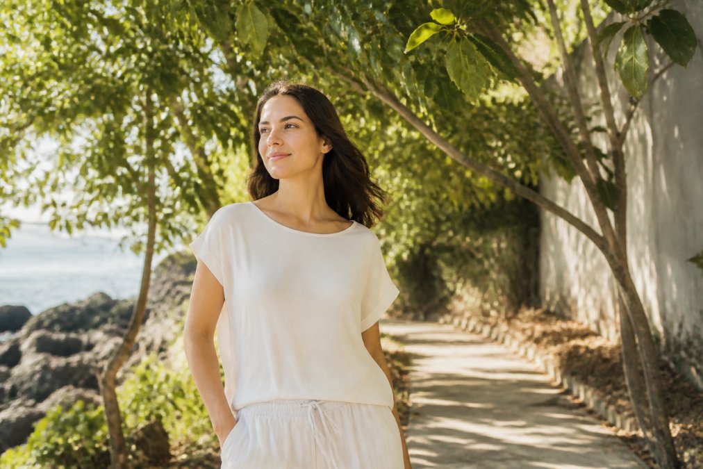 A woman in a loose, light-colored bamboo fabric top standing outdoors on a sunny afternoon, looking relaxed with a gentle breeze, soft natural light filtering through nearby trees