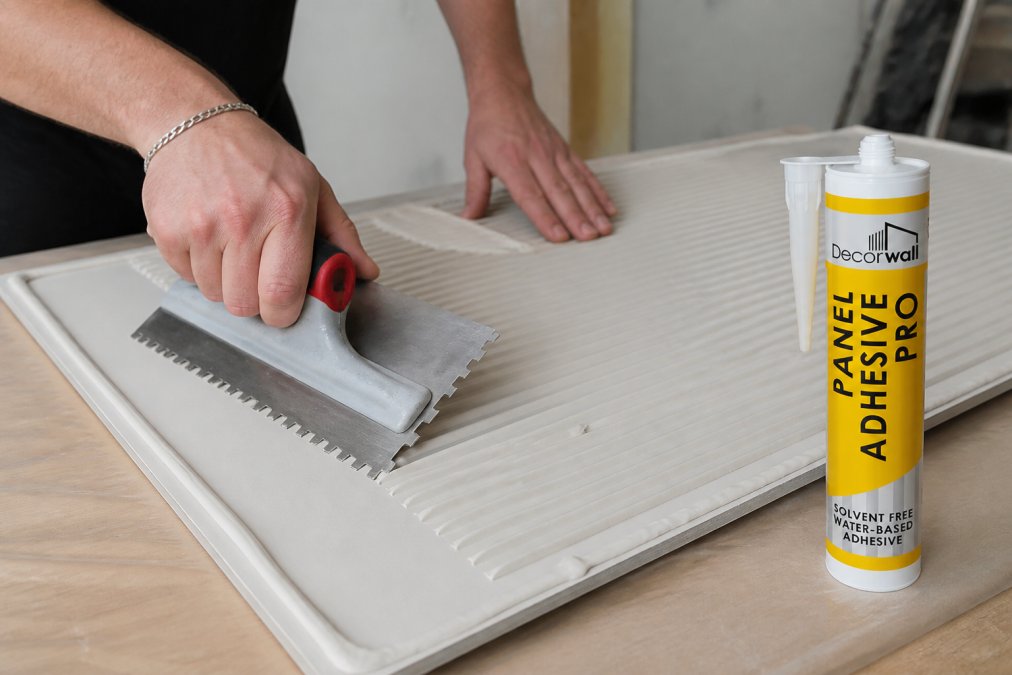 Close-up of hands applying adhesive to the back of a bathroom wall panel using a notched spreader, showing consistent adhesive ridges and proper technique with beads visible