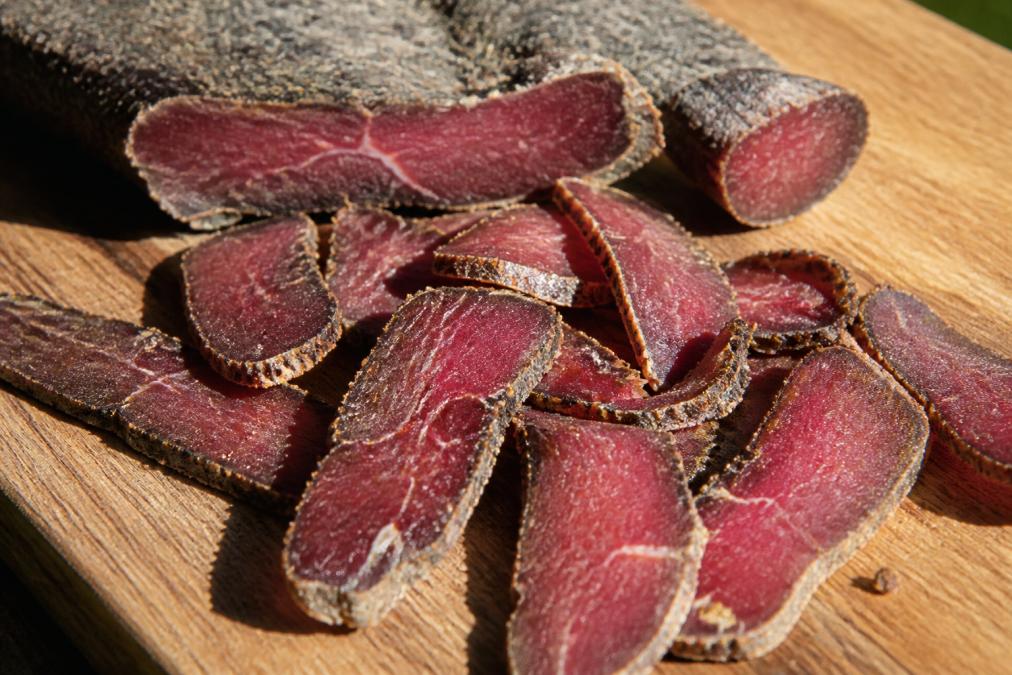 Close-up of sliced biltong on a wooden board with visible marbling and texture, natural sunlight highlighting the deep red-brown color of the cured meat
