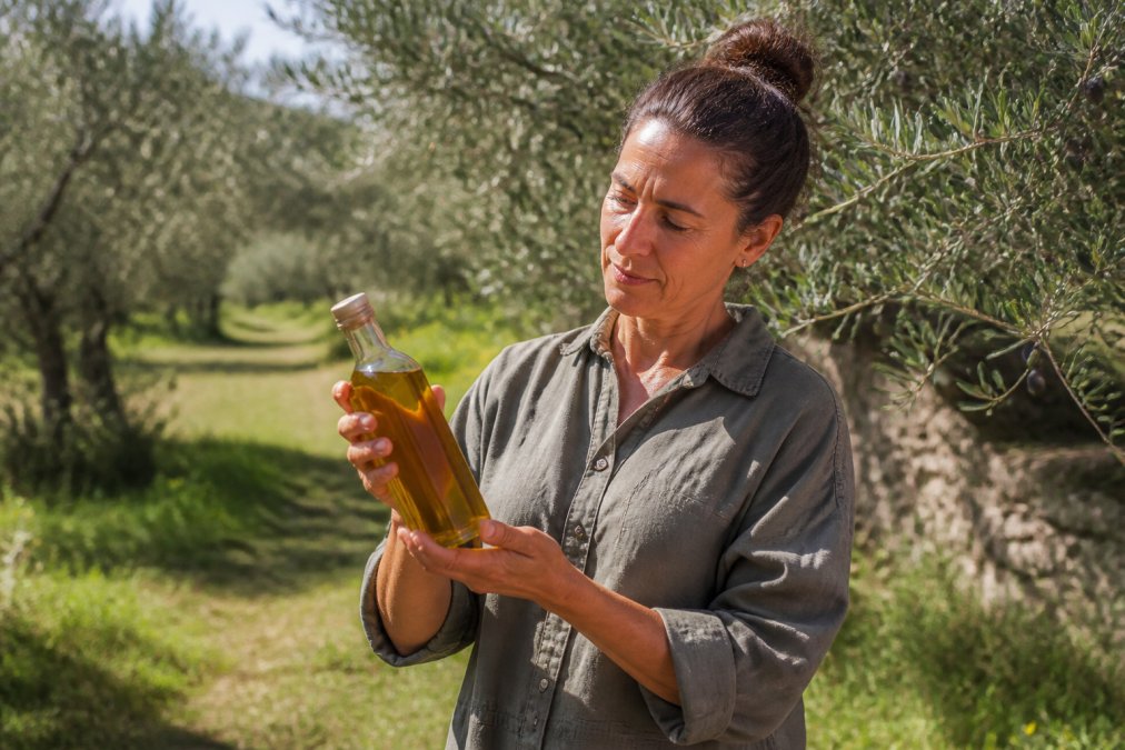 Eén persoon in een zonnige olijfgaard die een glazen fles biologische olijfolie vasthoudt, met olijfbomen en natuurlijke begroeiing op de achtergrond, warm natuurlijk licht