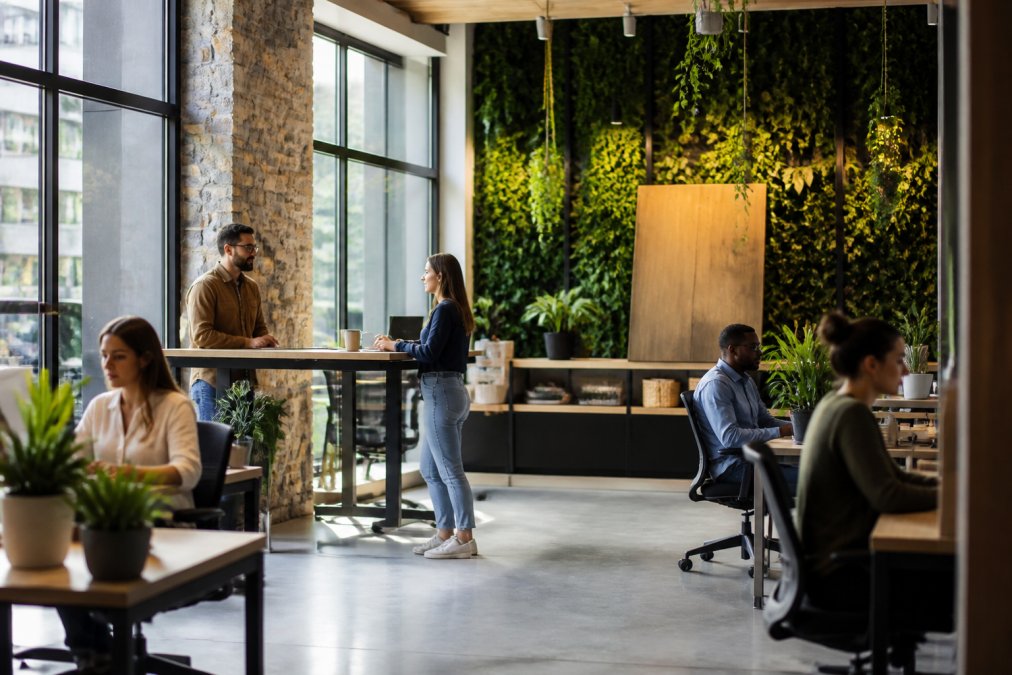 Modern office workspace with abundant natural light streaming through large windows, living green walls with cascading ivy, potted plants on desks and shelves, employees working at standing desks surrounded by natural wood materials and stone accents, bright and airy environment