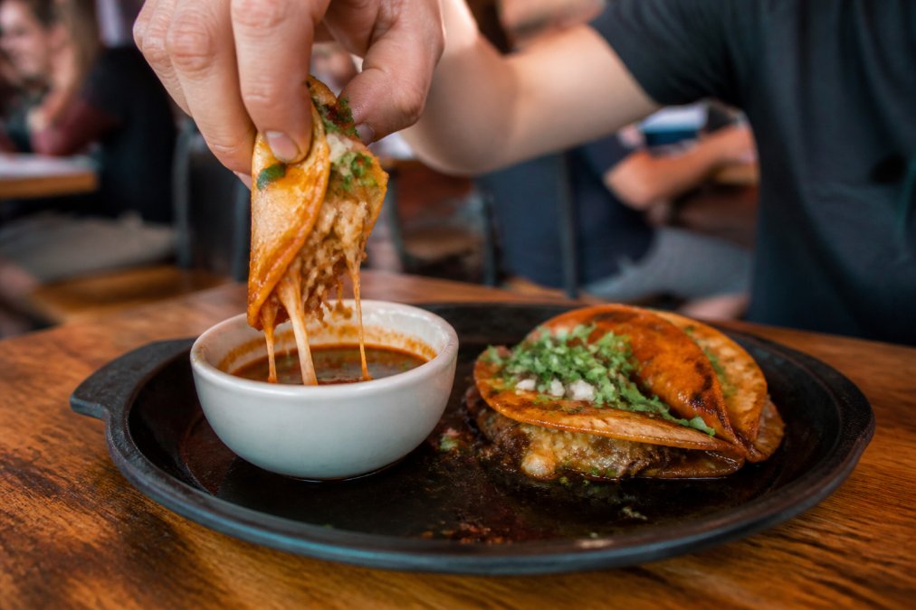 Close-up of two birria tacos on a cast iron plate, one being dipped into a small bowl of red consommé, with melted cheese stretching from the tortilla under warm restaurant lighting