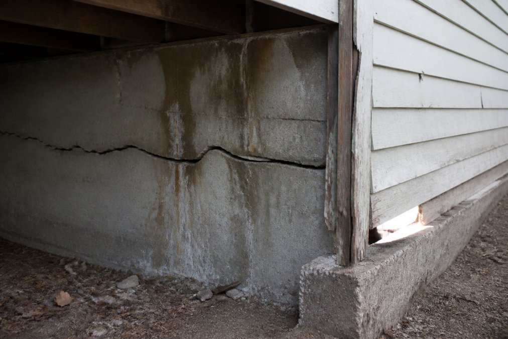 Close-up of visible horizontal foundation cracks with water staining on basement concrete wall, daylight visible through gaps in older home exterior