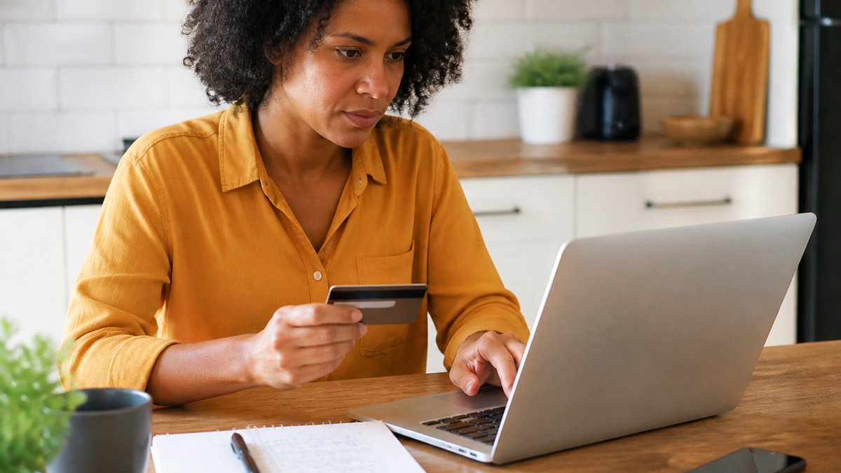 A person sitting at a kitchen table with a laptop open to an online store page, one hand on a credit card, a notebook with handwritten checkmarks beside the keyboard