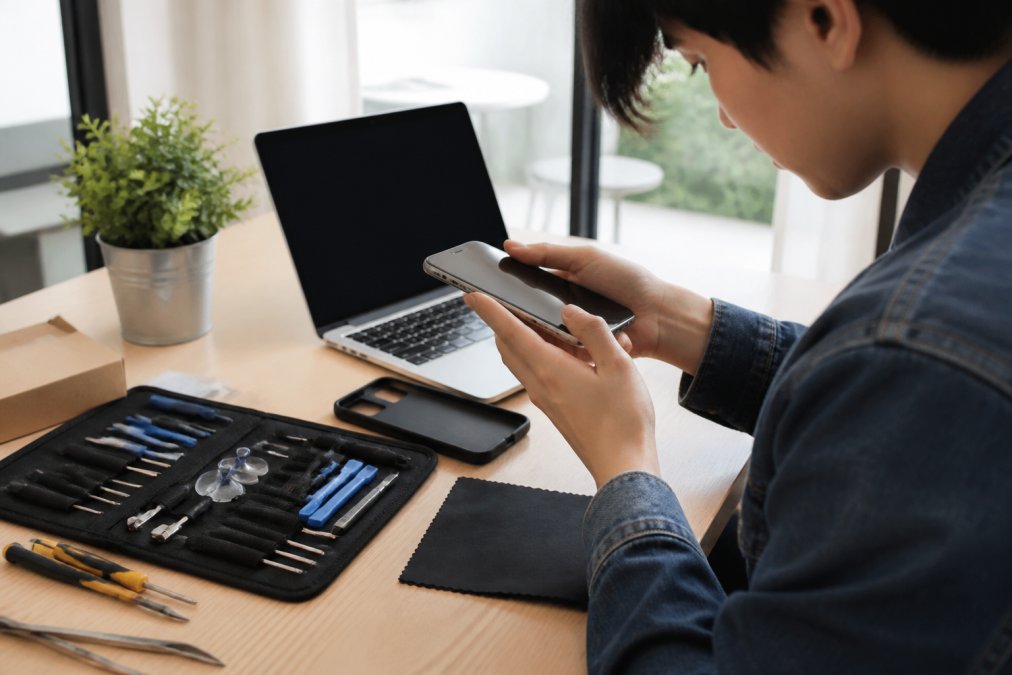 A young person in a bright home office inspecting a refurbished smartphone under natural light, checking the screen and ports with a laptop open nearby showing a product listing page