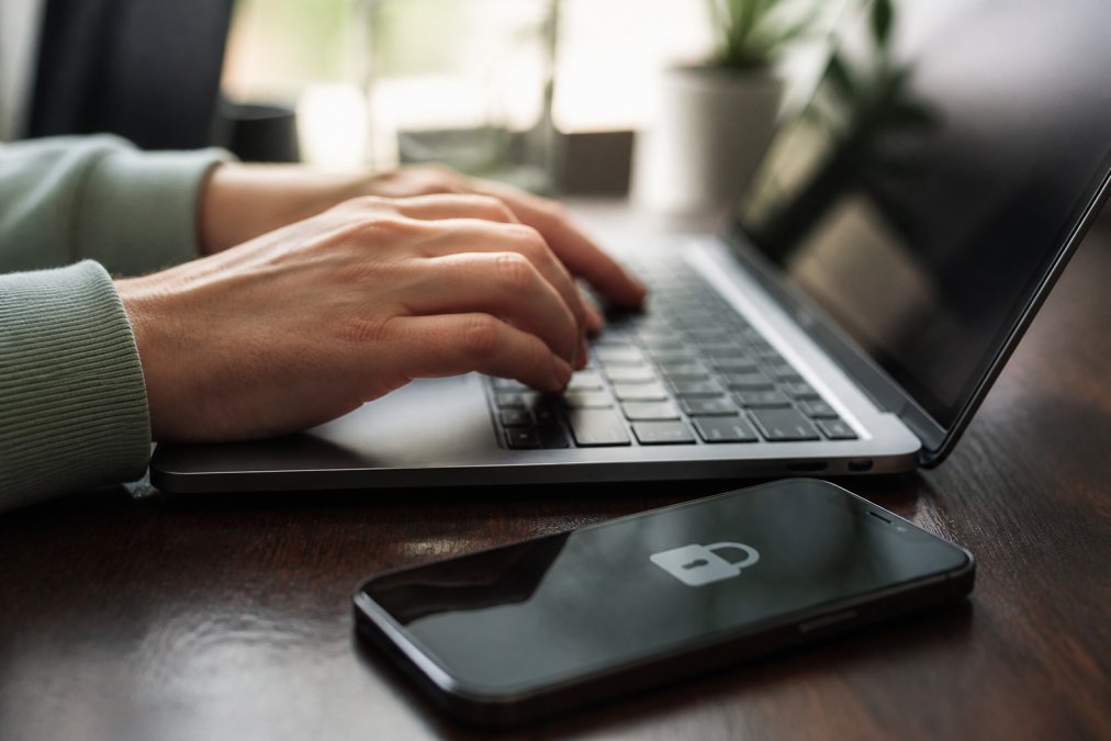 Close-up of hands typing on a laptop keyboard with a smartphone nearby showing a padlock security icon, representing secure online shopping