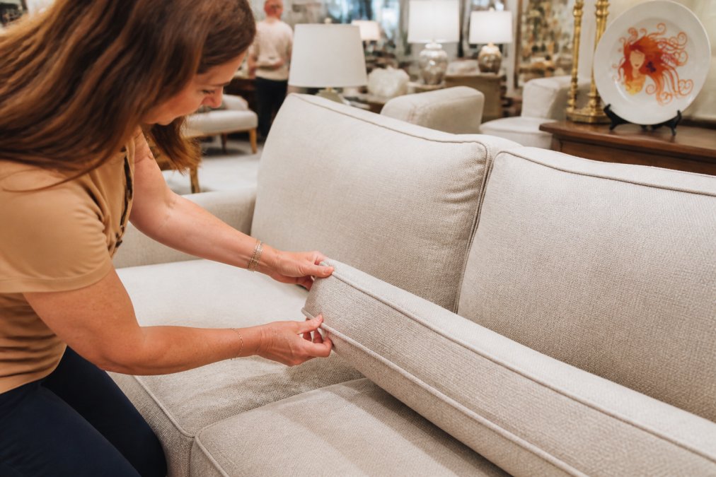 Close-up of hands inspecting upholstery seams and checking for stains and wear patterns on a used sofa cushion in a bright consignment store showroom