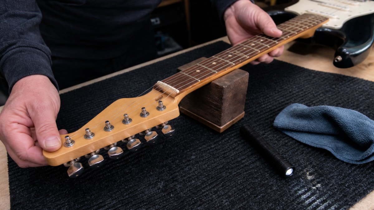 Close-up of a person's hands turning a tuning peg and inspecting the neck of a second-hand electric guitar on a workbench, with a small torch and cloth nearby