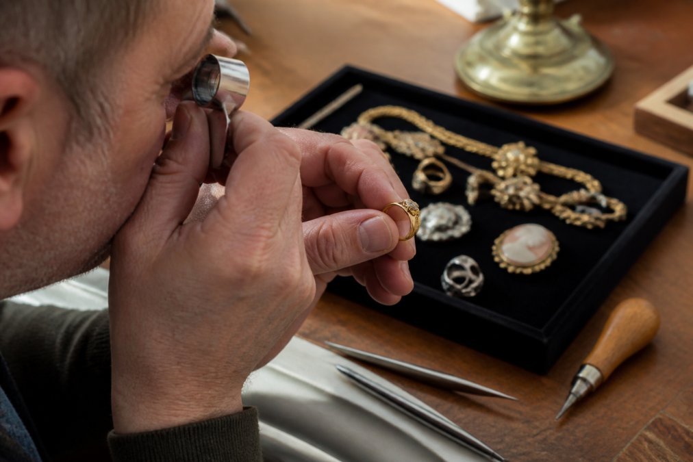 Close-up of a jeweler's hands using a loupe to examine hallmarks on a vintage gold ring resting on a black velvet display tray, warm natural lighting from a desk lamp illuminating the gold surface