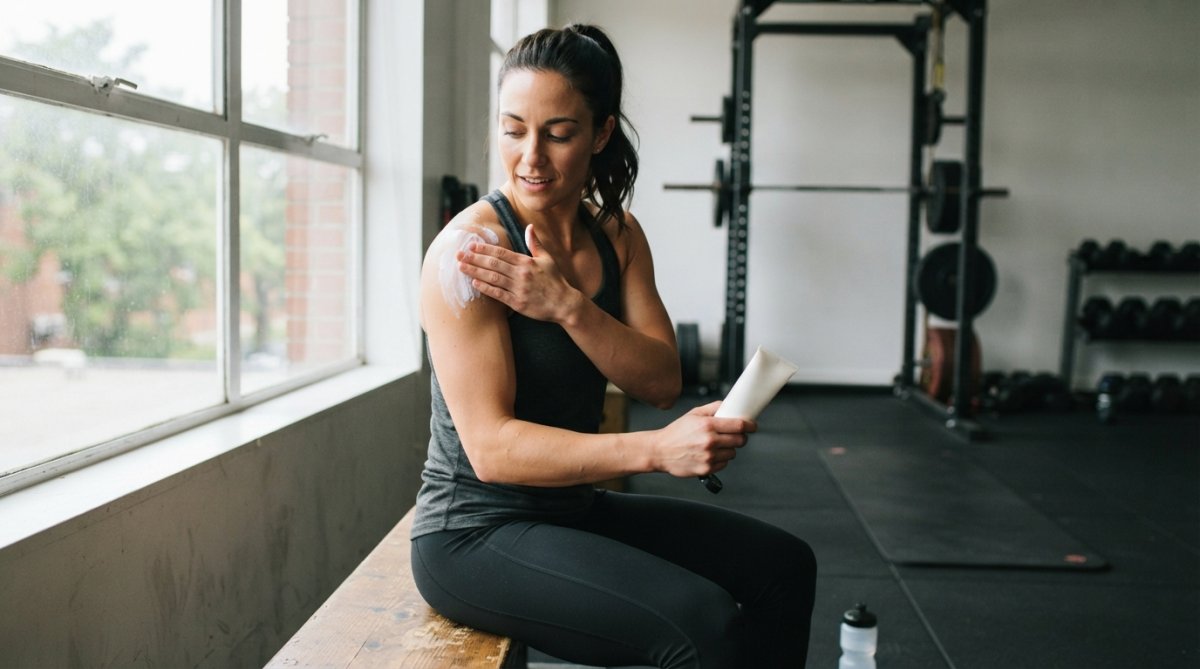 Athlete applying cream to shoulder after workout in gym setting with equipment visible in background and natural window lighting