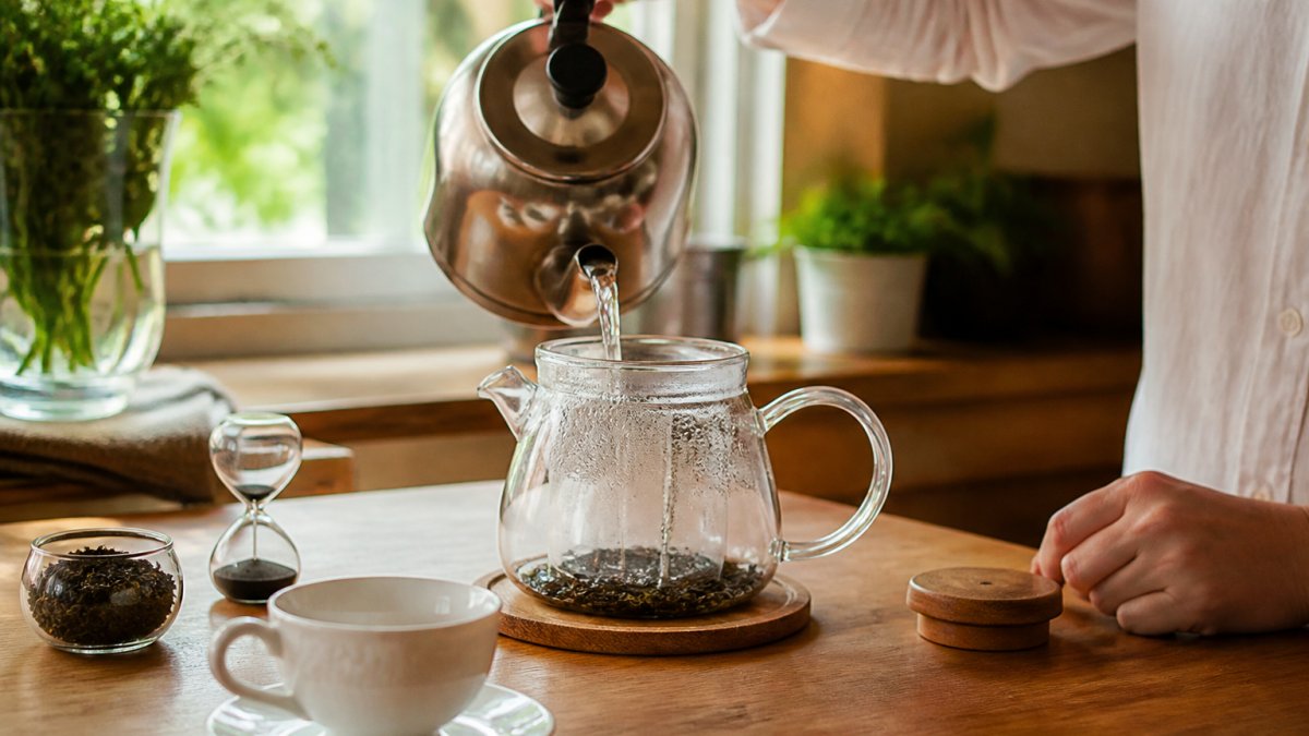 Person pouring hot water over loose tea leaves in a clear teapot on a sunny kitchen counter