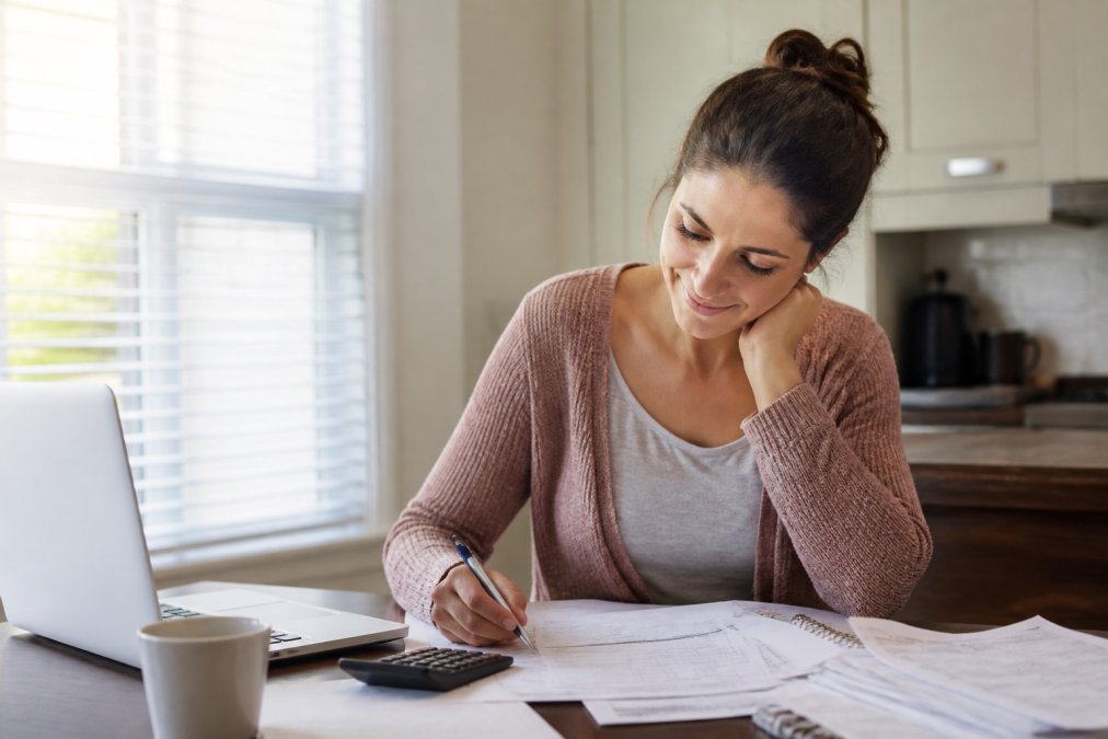 A person sitting at a kitchen table with a laptop and a stack of bills, looking relieved while reviewing a financial plan on paper, soft morning light through a window