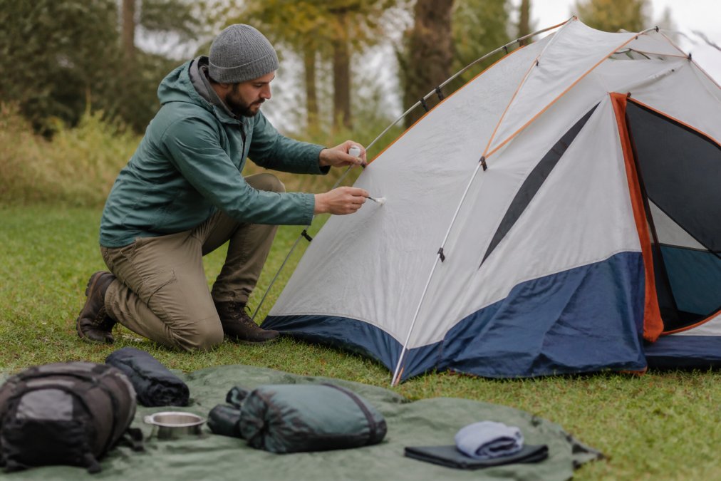 A camper kneeling beside a pitched dome tent in a grassy campsite, applying seam sealant to the tent's rainfly with a small brush, camping gear laid out on a groundsheet nearby