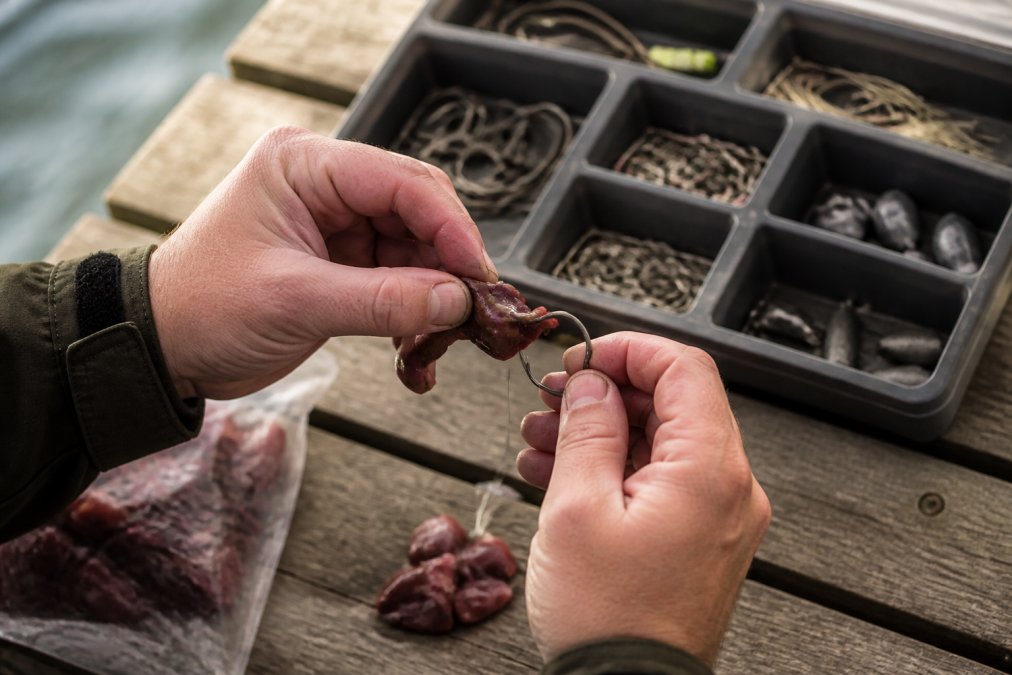 Close-up of hands rigging a piece of chicken liver onto a circle hook, with a tackle box containing various hooks and terminal tackle visible in the background on a wooden dock