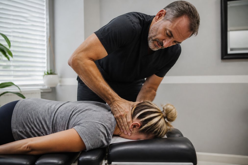 Licensed chiropractor performing cervical spine adjustment on patient lying face-down on treatment table, hands positioned carefully on neck area with focused concentration, modern clinical setting with soft overhead lighting
