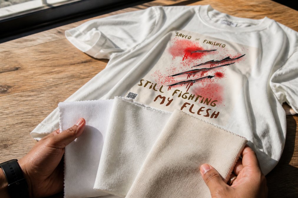 Close-up of hands holding and examining different fabric swatches, 100% cotton, cotton-polyester blend, and heavyweight jersey, showing texture and weight differences under natural window lighting